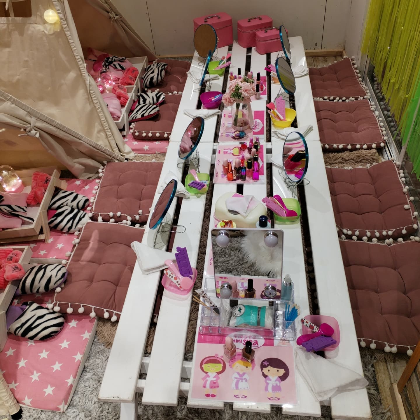 Mesa de spa infantil con espejos, esmaltes, cintillos y cojines rosados alrededor