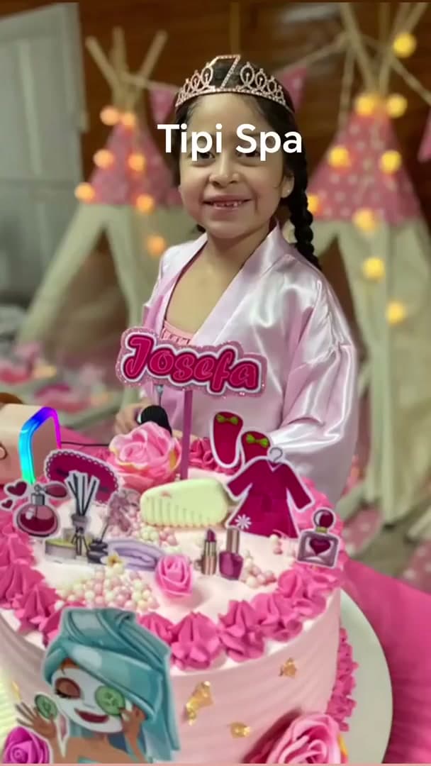 Niña con bata rosada y tiara frente a su torta temática de spa, con tipis iluminados de fondo