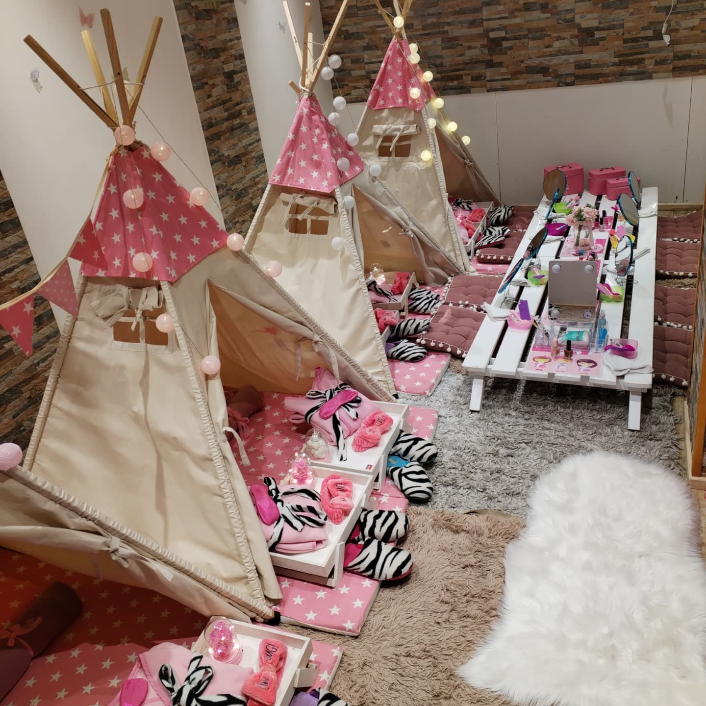 Tres carpas tipi con estrellas rosadas, guirnalda de luces, colchonetas con cojines de cebra y mesa de spa al fondo